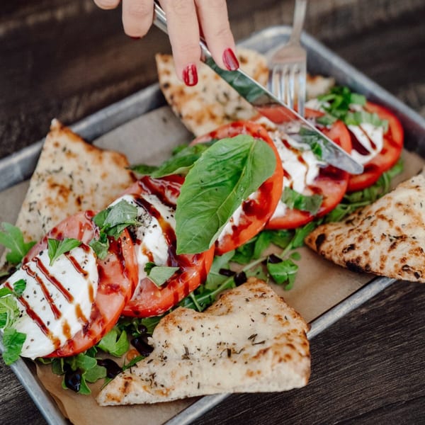 A person cuts into a fresh Caprese salad with basil, tomatoes, mozzarella, and balsamic glaze, served with pizza bread.