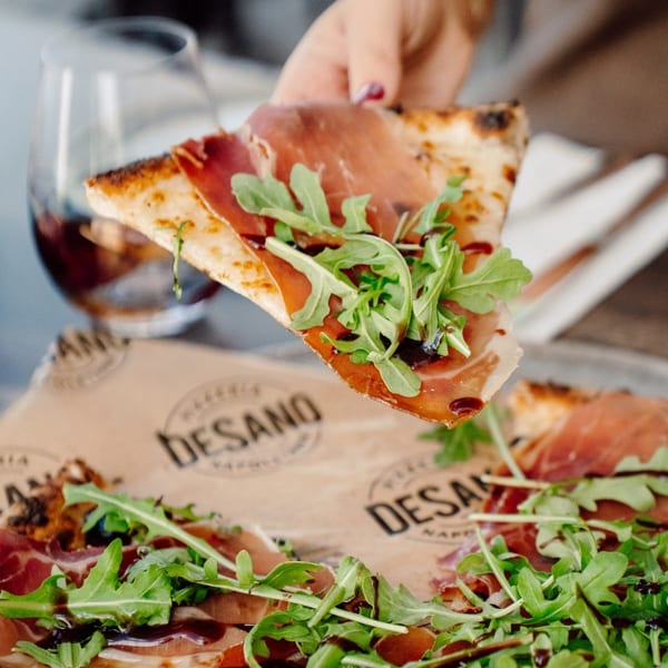 Hand holding a slice of DeSano Pizzeria Napoletana pizza topped with prosciutto, arugula, and balsamic glaze.
