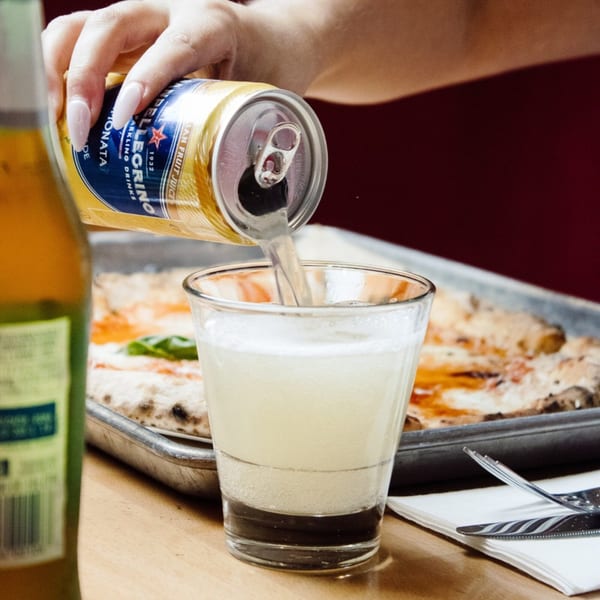 A hand pours a San Pellegrino sparkling drink into a glass, with a pizza in the background.