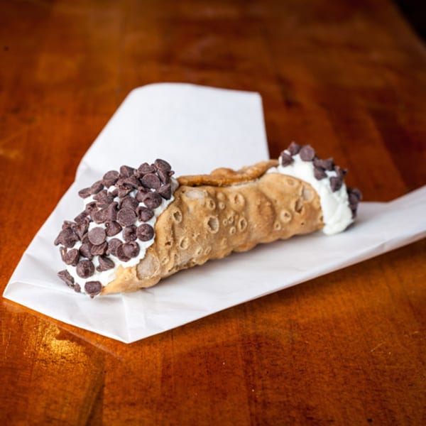 A DeSano Pizzeria Napoletana cannoli, filled with cream and topped with chocolate chips, on white paper.