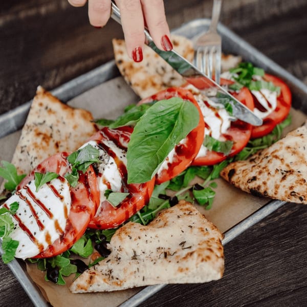 Hand cutting a Caprese salad with fresh mozzarella, tomatoes, basil, and flatbread on a tray.